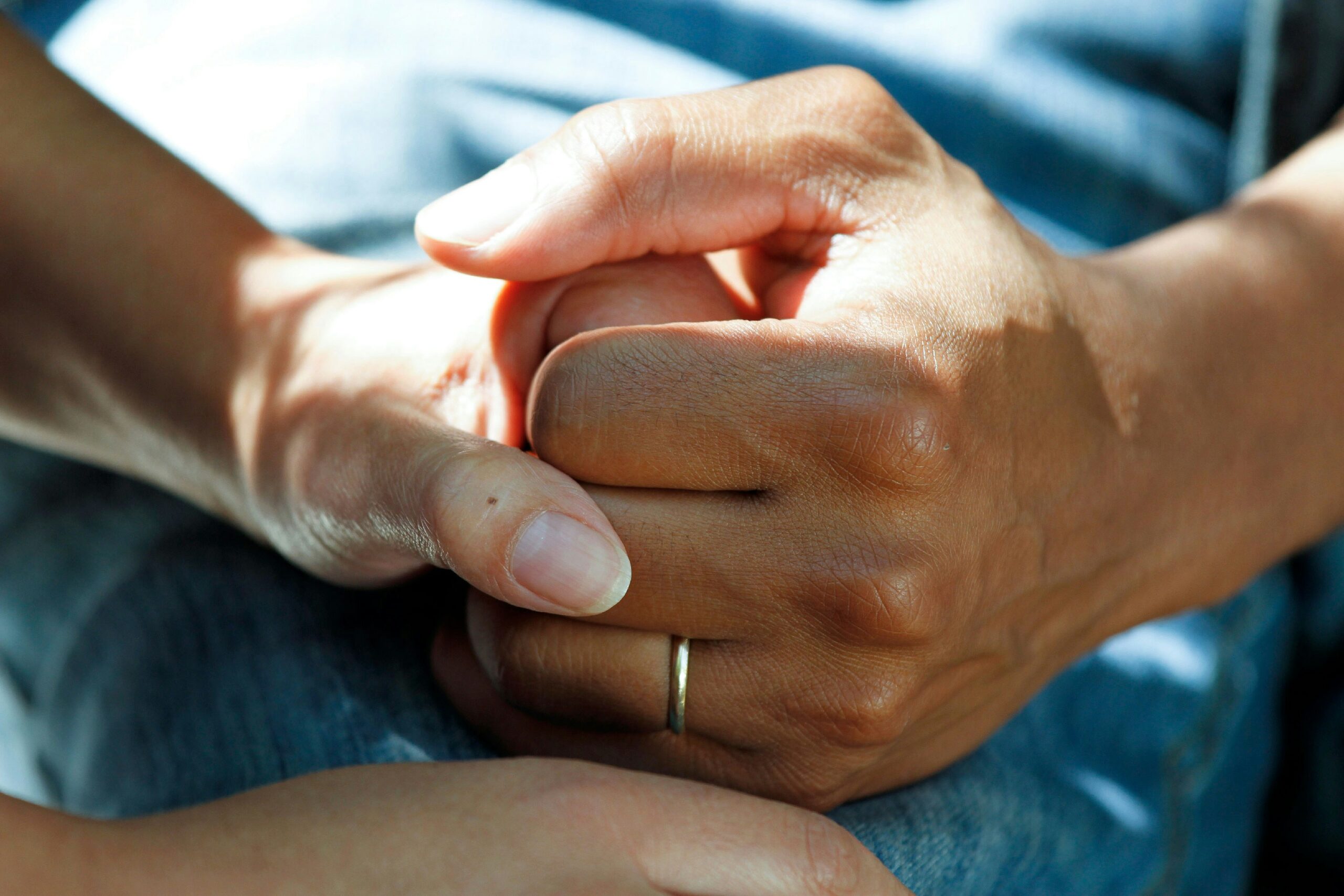 Close-up of two hands gently clasped together, with a silver ring on one finger and blue fabric in the background.