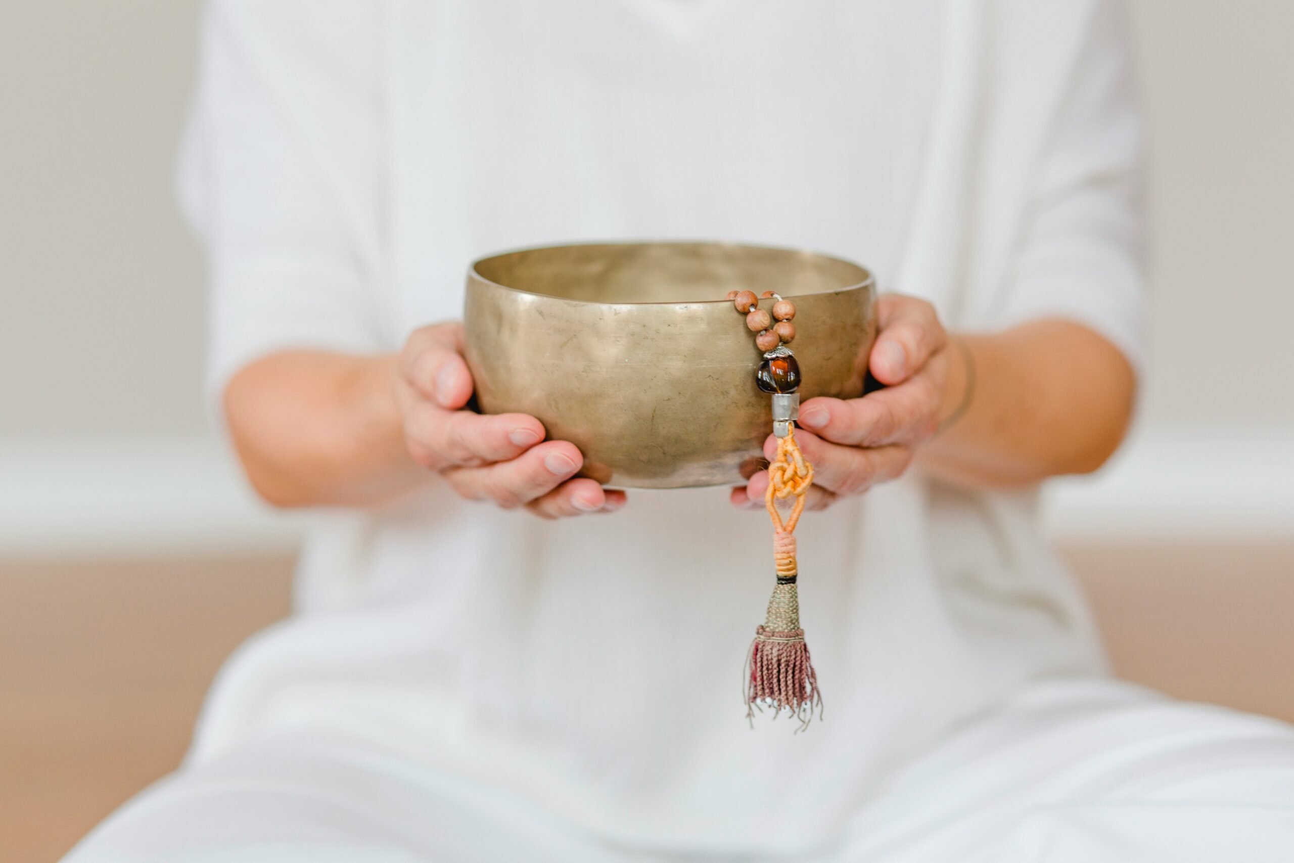 Person in white holds a brass singing bowl with a beaded bracelet and tassel hanging from its rim.